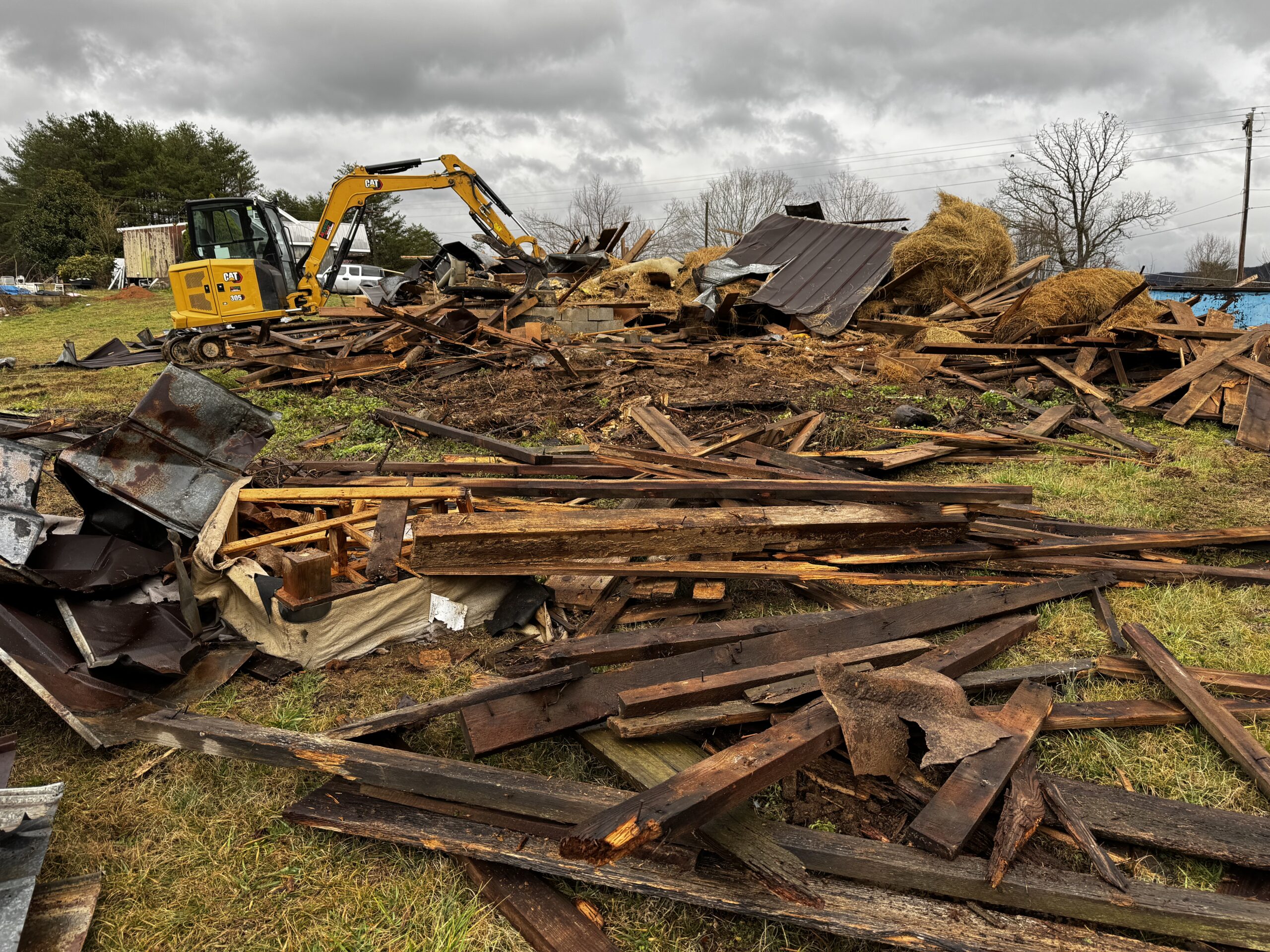 A yellow excavator sits beside a large pile of wooden debris and metal scraps on a grassy field under a cloudy sky.
