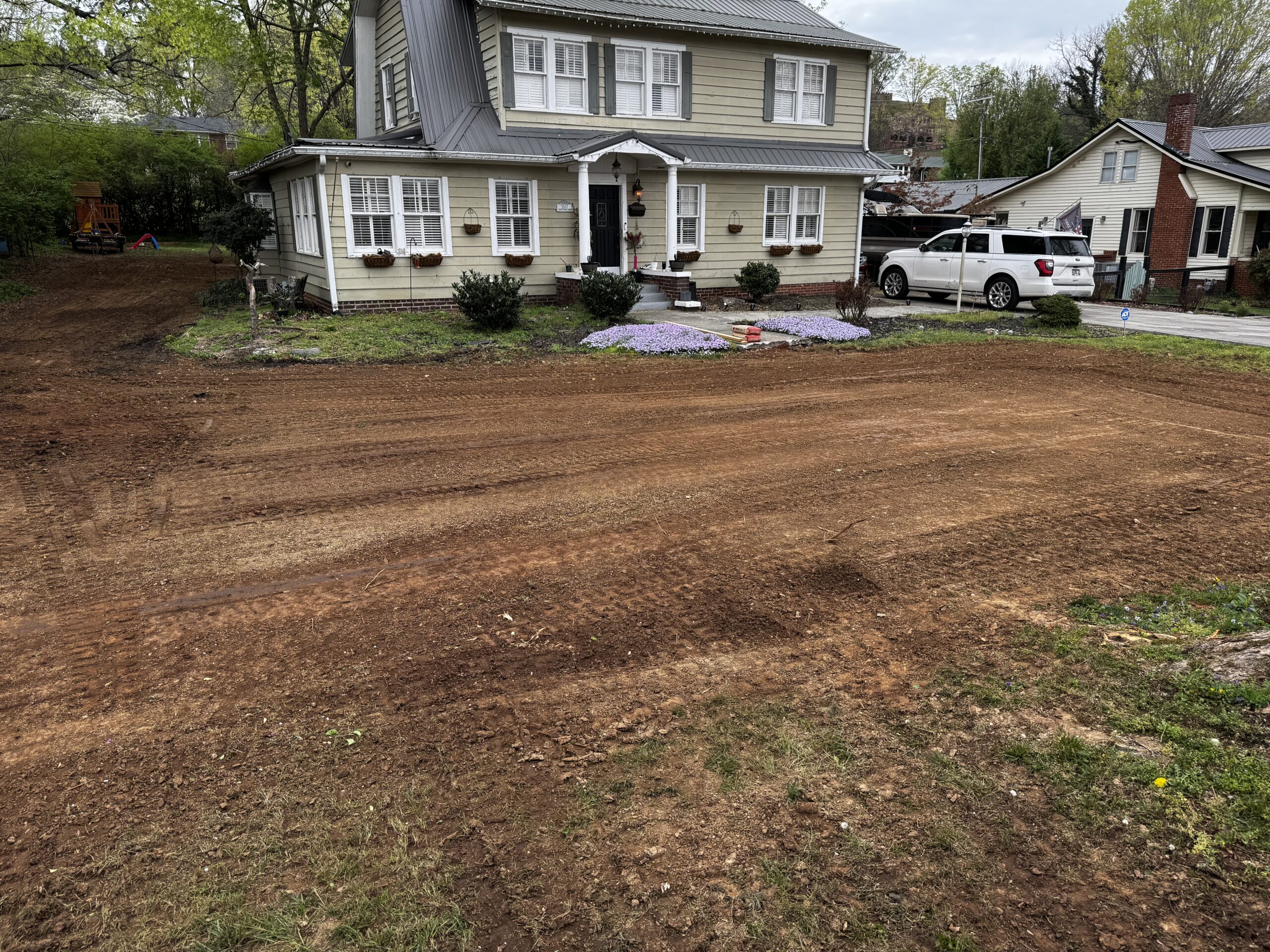 A house with a freshly graded dirt yard in front, small flower bed near the entrance, and a white SUV parked in the driveway.