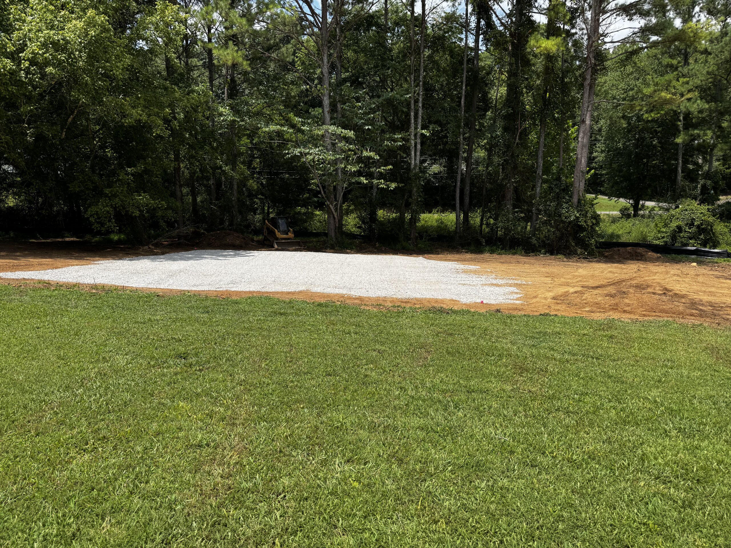 A patch of gravel spread on leveled ground near a grassy lawn, with trees and forested area in the background.