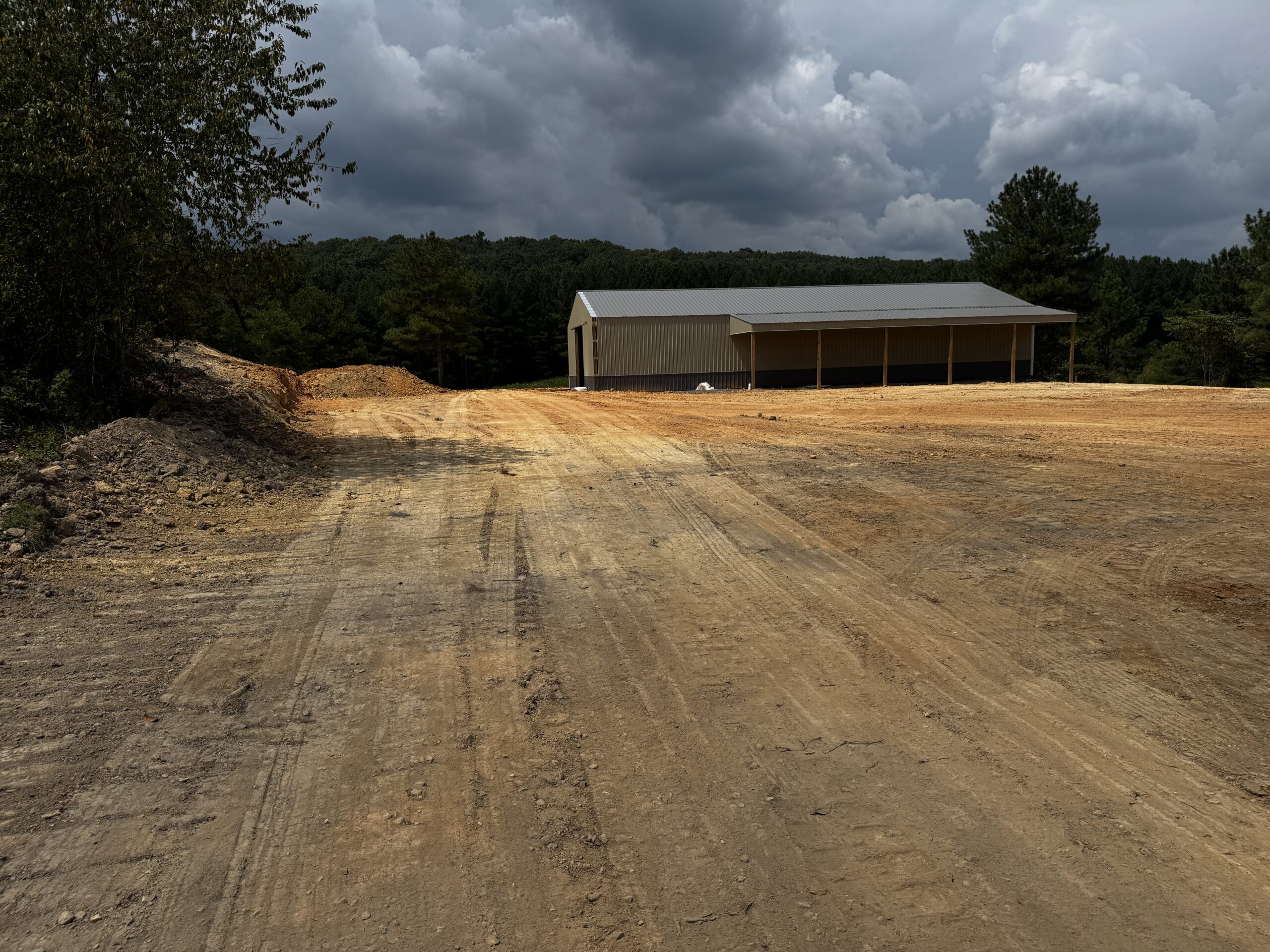 A large metal shed sits at the edge of a cleared dirt lot with trees and dark clouds in the background.