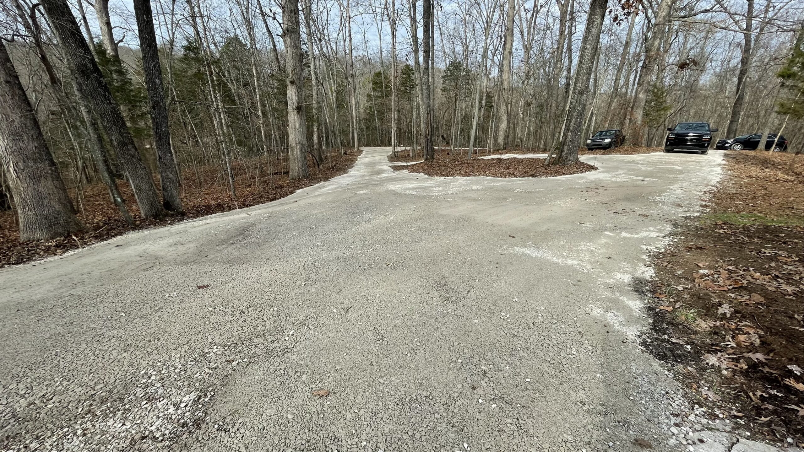 A gravel driveway curves through a wooded area with three parked vehicles visible on the right side among bare trees.