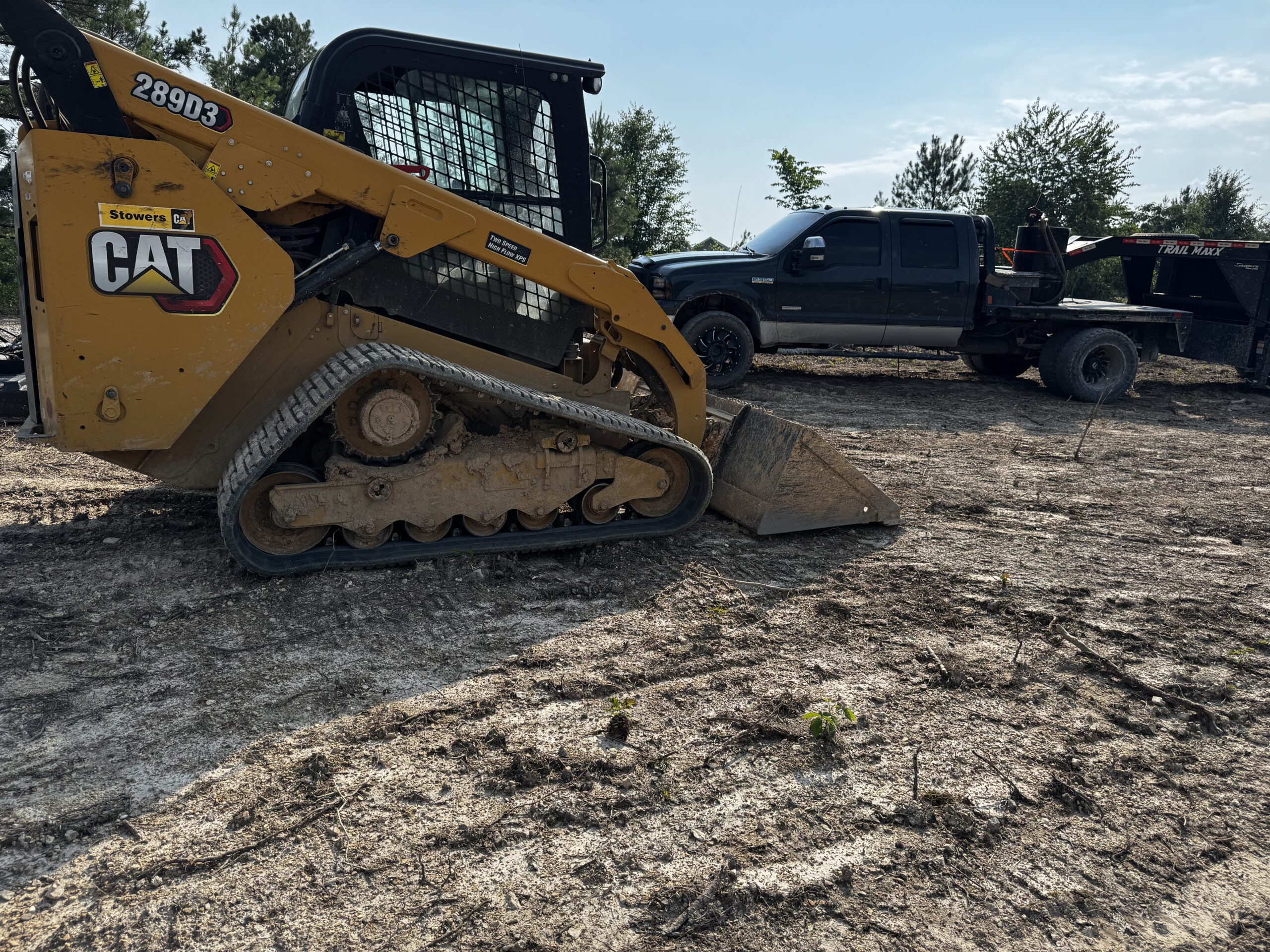 A yellow CAT compact track loader is parked on dirt near a black pickup truck and a flatbed trailer on a sunny day. Trees are visible in the background.