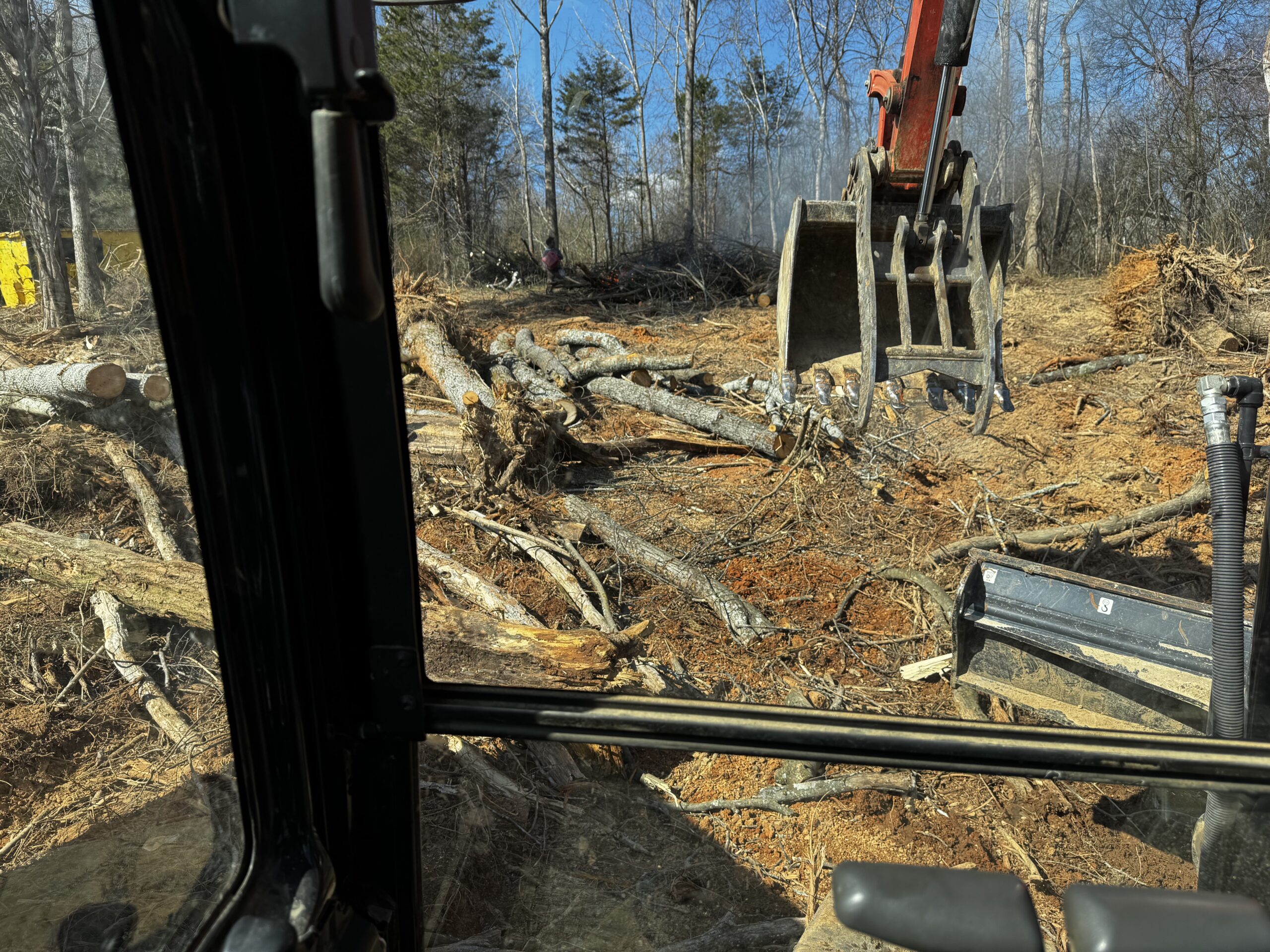 View from inside a vehicle cab showing an excavator clearing tree trunks and debris from a forested area on a sunny day.