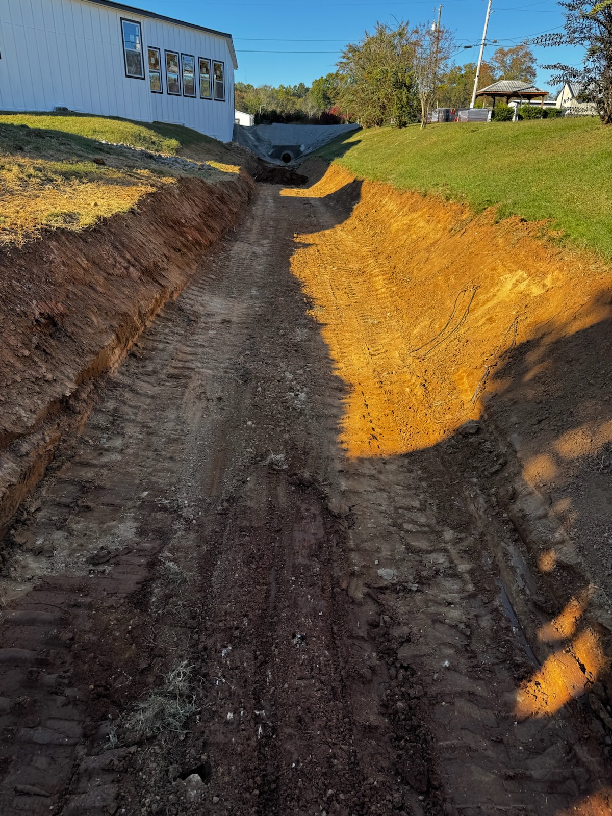 A freshly excavated dirt trench by an experienced excavating contractor runs alongside a building, bordered by grass and utility poles beneath a clear sky.