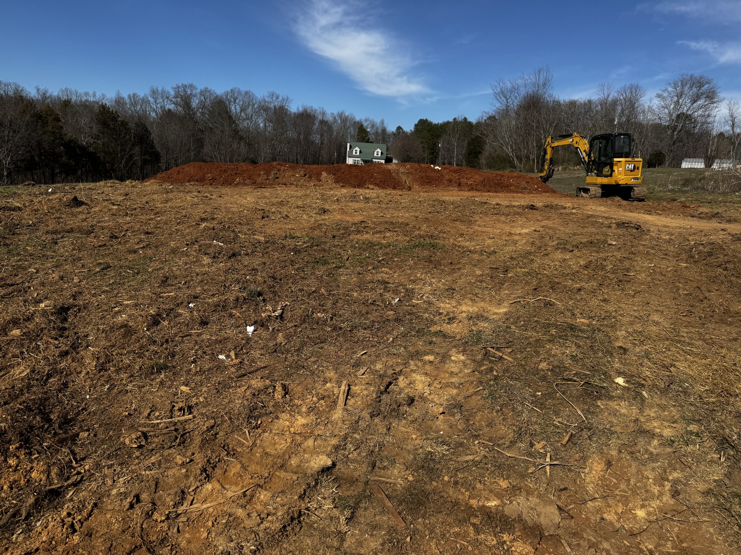 A construction site with cleared dirt, an excavator on the right, and a pile of soil; trees and a house with green roof in the background under a blue sky.