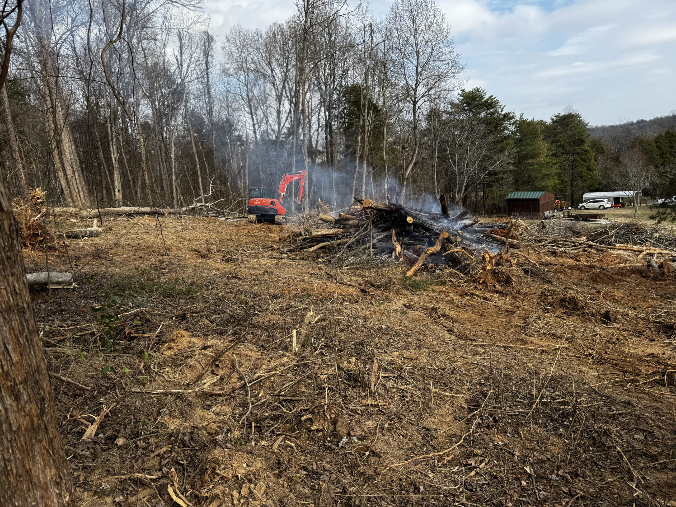Cleared forest area with a pile of burning logs and smoke, a red excavator, and vehicles by a small building in the background.