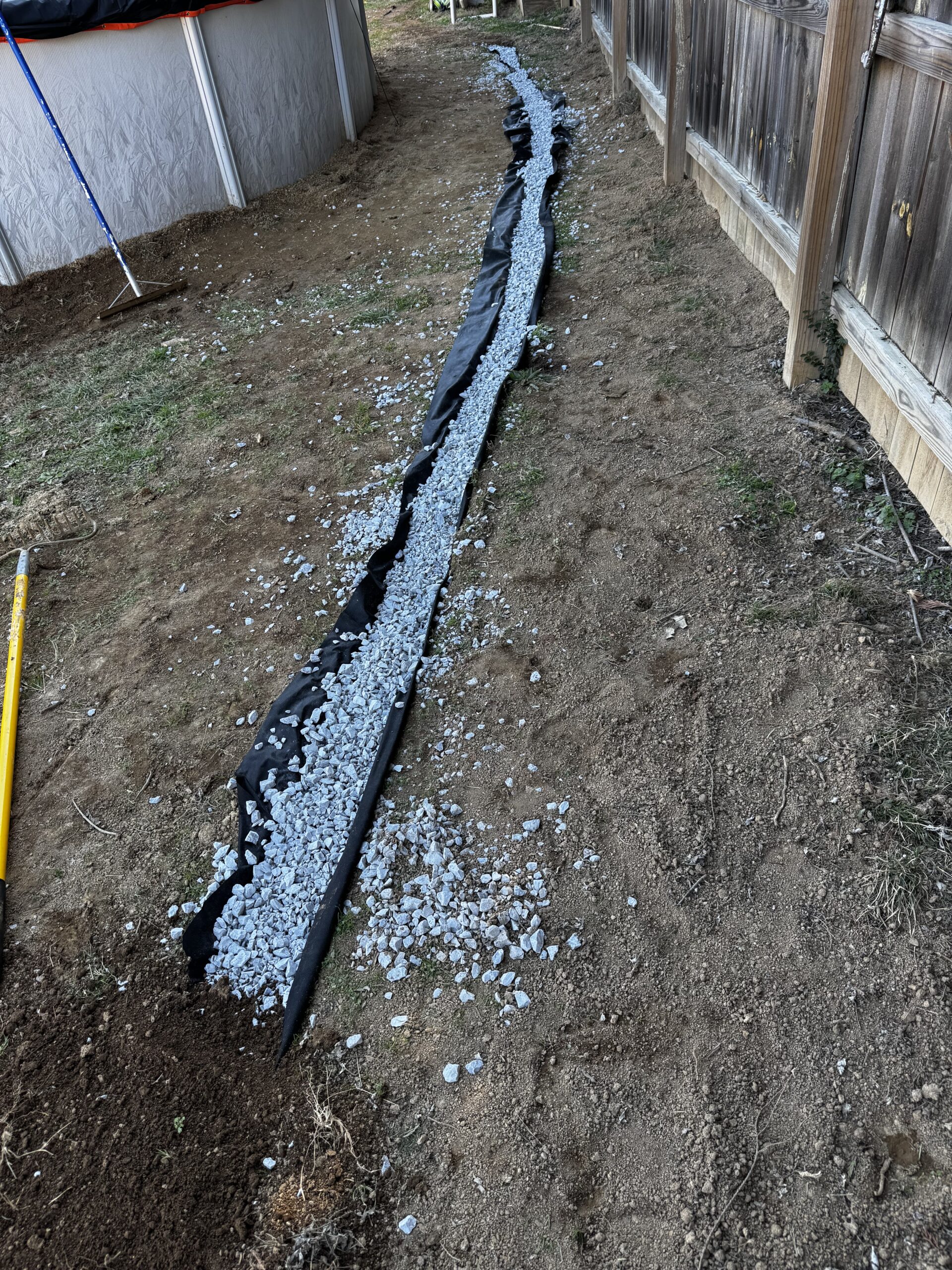 A narrow trench lined with black landscape fabric and filled with gray gravel runs along a wooden fence in a backyard.
