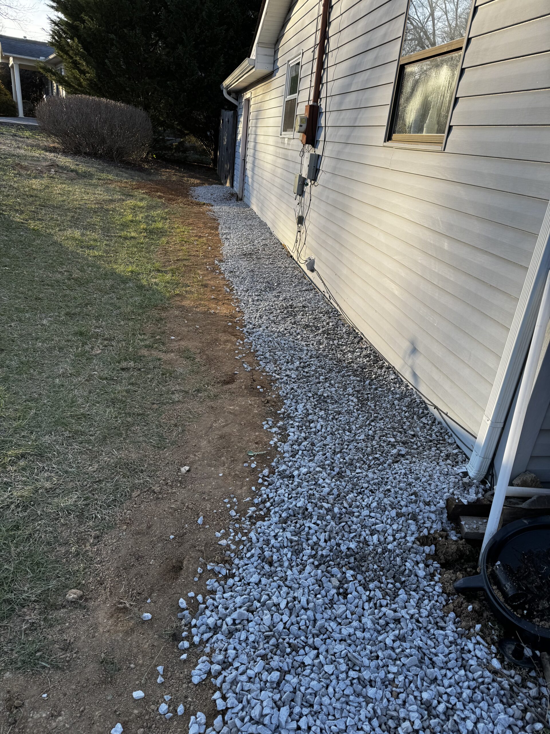 A layer of gravel is spread along the side of a beige house, separating the building from the adjacent grass and dirt area.